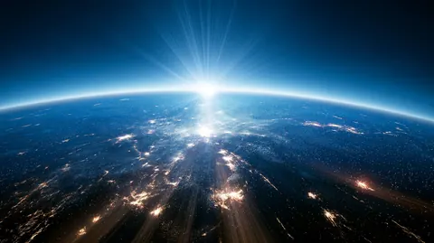 The sun rises over the curve of the Earth, lighting the city lights scattered across the night side.